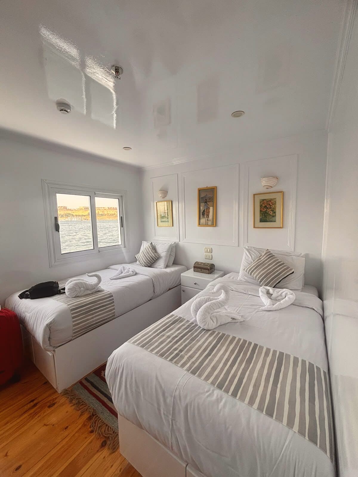 Cabina de dos camas individuales con ropa blanca, piso de madera y ventana con vista a la orilla del río