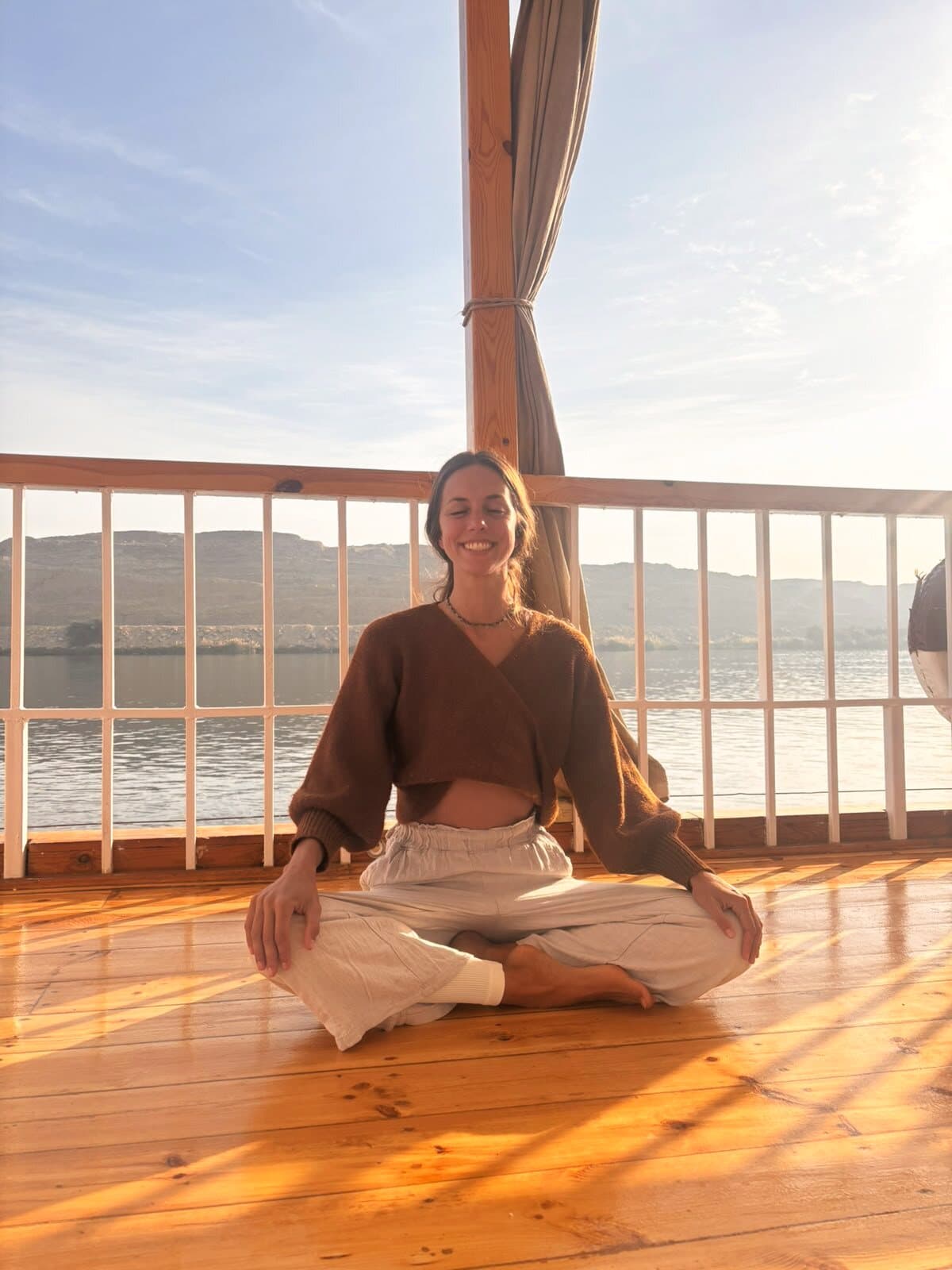 Mujer sentada en meditación sobre la cubierta de madera, con el sol bajo y las montañas detrás