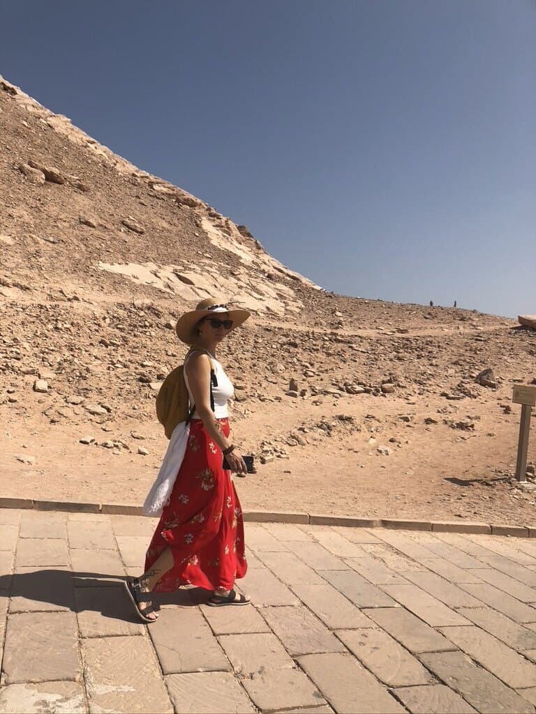 Mujer con sombrero de ala ancha caminando sola por un sendero de piedra al pie de una ladera de arena