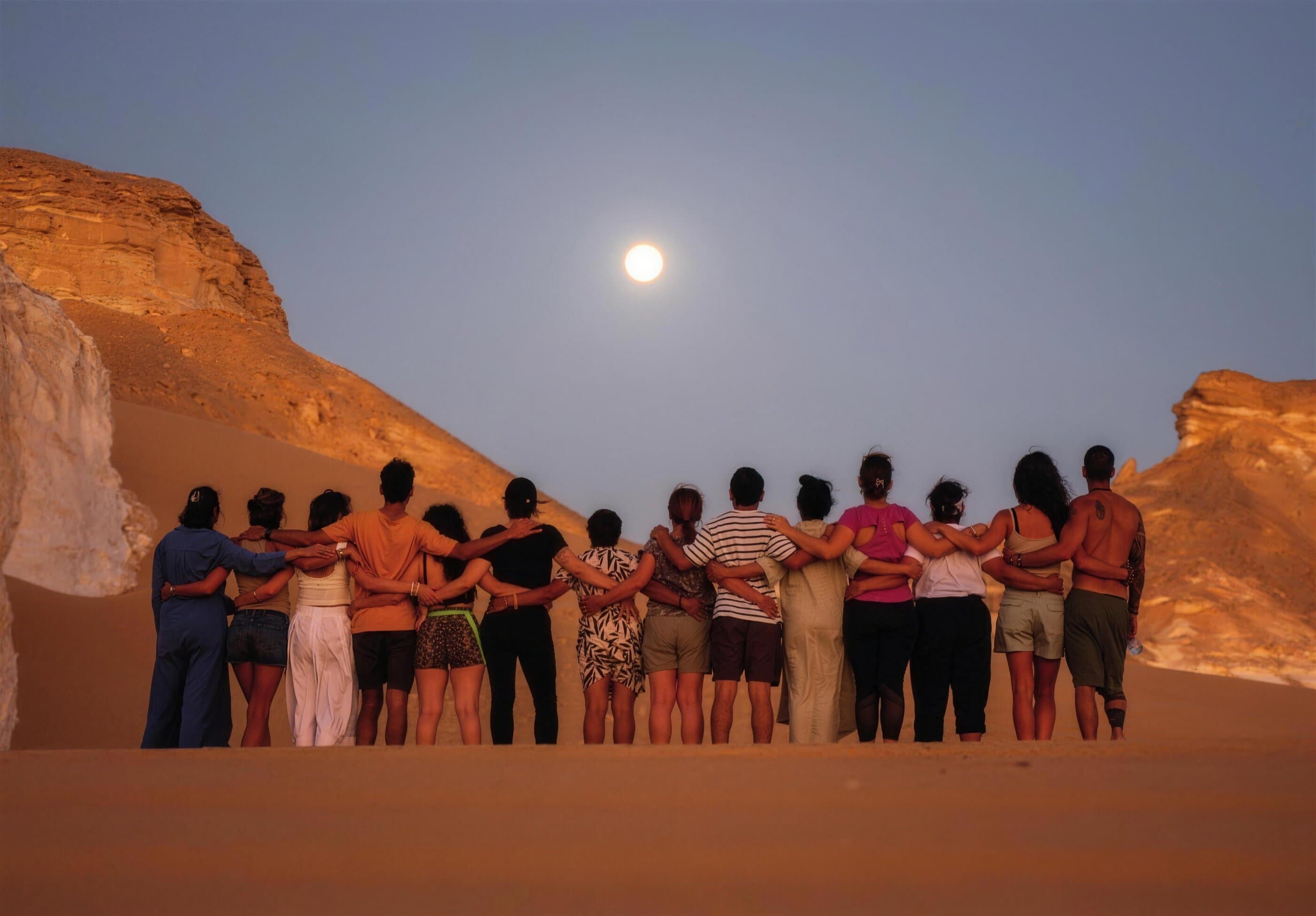 El grupo entero abrazado de espaldas sobre una duna del desierto, con la luna llena saliendo sobre las rocas