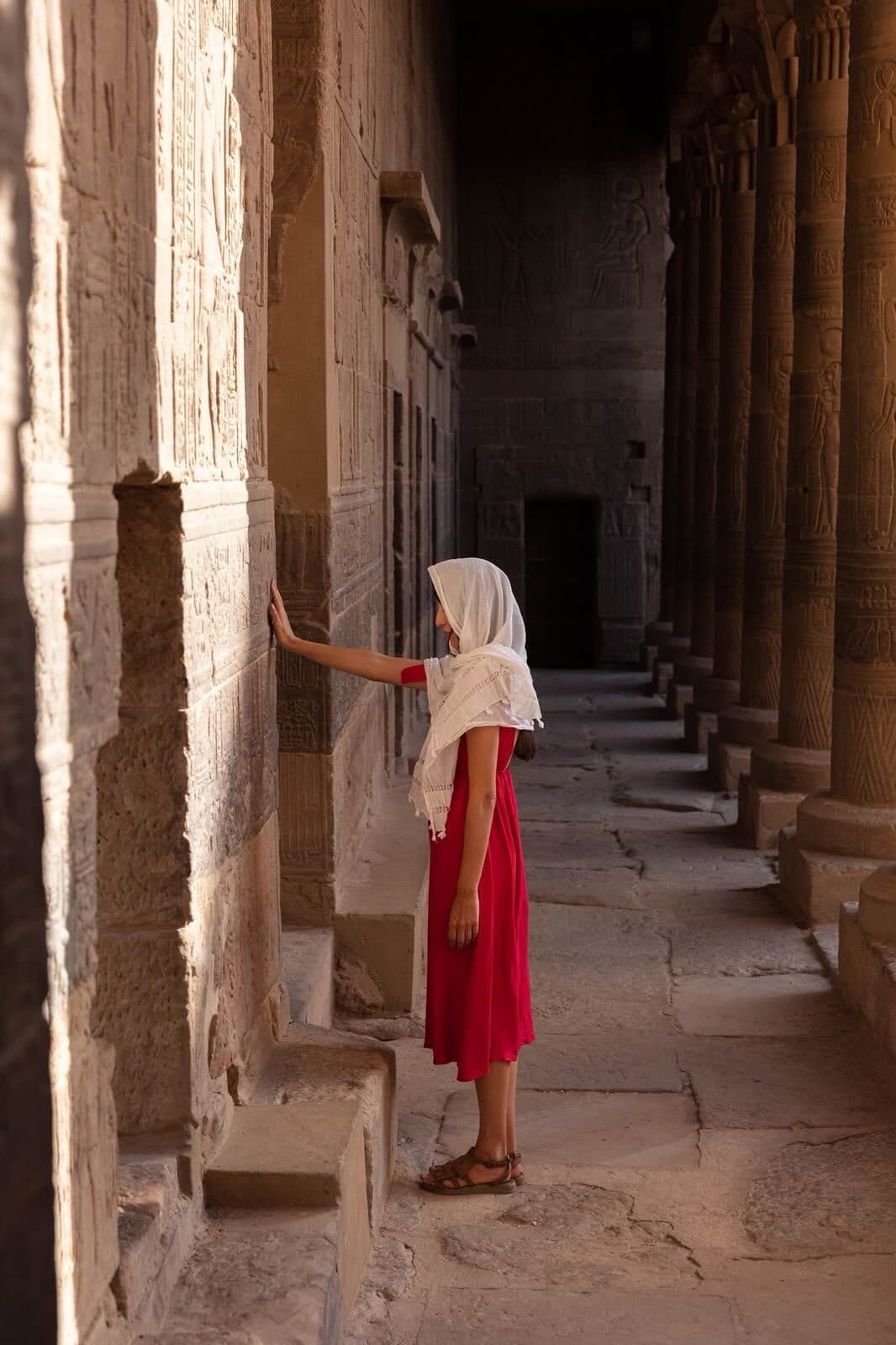 Viajera de rojo con velo blanco apoyando la mano en una columna del templo